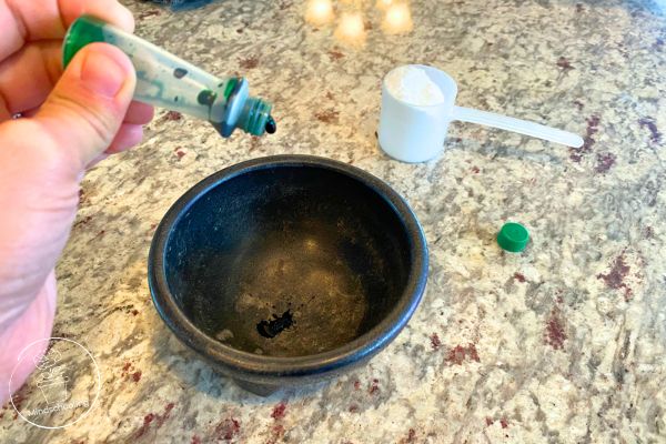 Someone prepping for the Harry Potter SEL Sorting Hat activity. There is green food coloring being dropped in a small black bowl with a scoop of baking soda sitting near by
