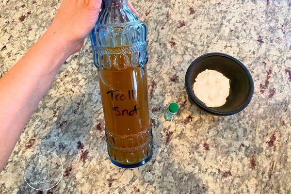 Someone prepping for the Harry Potter SEL Sorting Hat activity. They are holding a glass bottle filled with vinegar, with the label "Troll Snot" written on it. There is baking soda in a small black bowl with a few drops of green food coloring both in the bowl and the bottle sitting near by