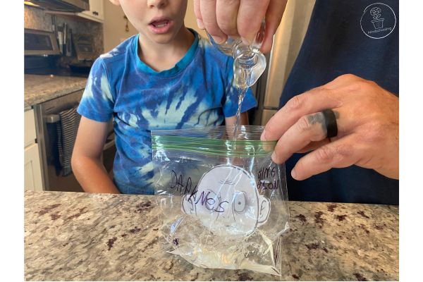 Harry Potter Defeat Voldemort Activity - There is a baggie with Voldemort's head from the printable taped on it and various fears written on the front. A child is sitting watching an adult pout vinegar into the baggie