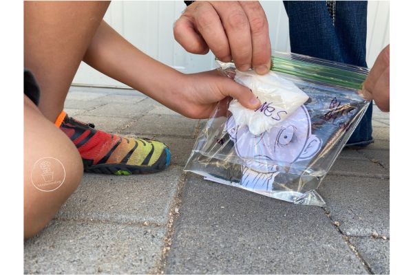This image is a close up of the baggie, with liquid inside, is being held by a child's hand, which is also holding the baking soda packet inside the bag but above the liquid. The adult's hand in the image is zipping the baggie closed